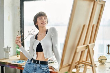 Woman standing, holding paintbrush in front of easel. clipart