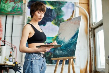 A woman in a black tank top is focused on painting on an easel. clipart