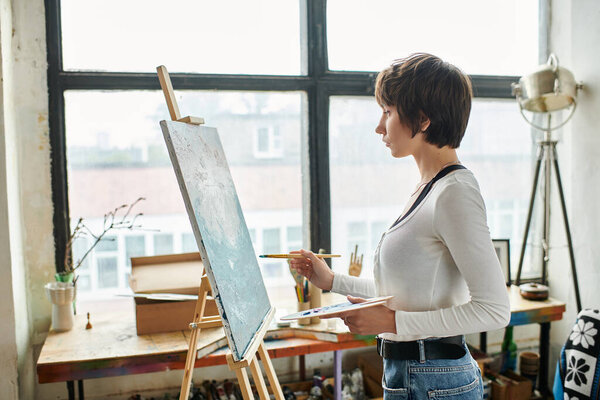 A woman stands confidently in front of a painting easel in an art studio.