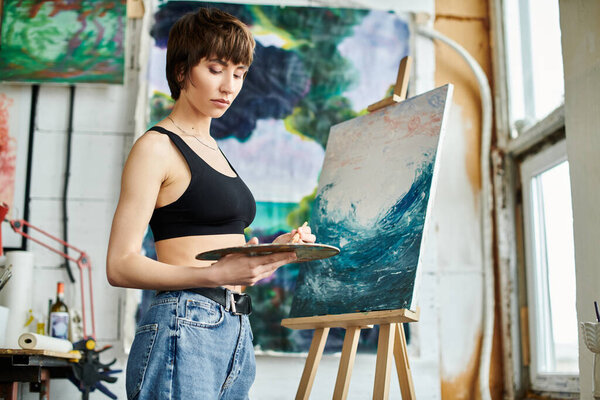 A woman in a black tank top is focused on painting on an easel.