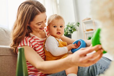 A young mother sitting on a couch, cradling her toddler daughter in her arms, sharing a special moment together. clipart