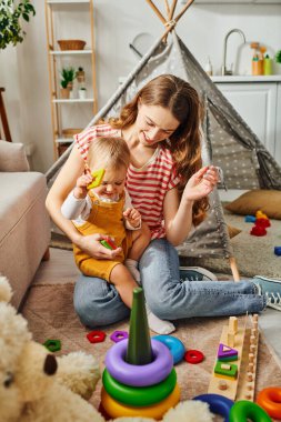 A young mother is happily playing with her toddler daughter in a cozy room, building a loving and joyful connection. clipart