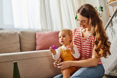 A young mother sitting on a couch, cradling her toddler daughter in her arms, sharing a moment of love and connection. clipart