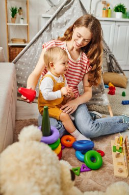 A young mother joyfully plays with her toddler daughter on the floor at home, bonding and creating happy memories together. clipart