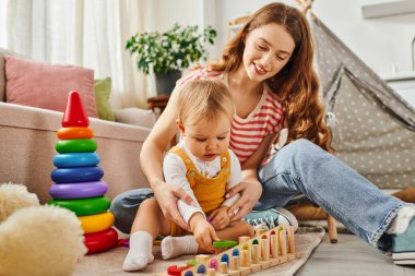 A young woman happily engages with her toddler daughter, playing on the floor in a home setting. clipart