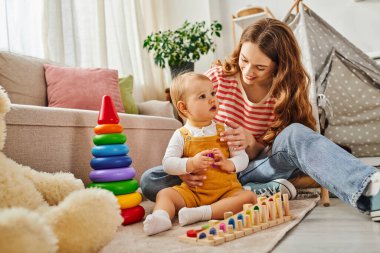 A young mother joyfully engages with her toddler daughter, playfully interacting in a warm and cozy living room setting. clipart