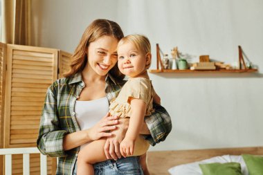 A young mother lovingly cradles her toddler daughter in her arms, creating a tender moment of connection and affection at home. clipart