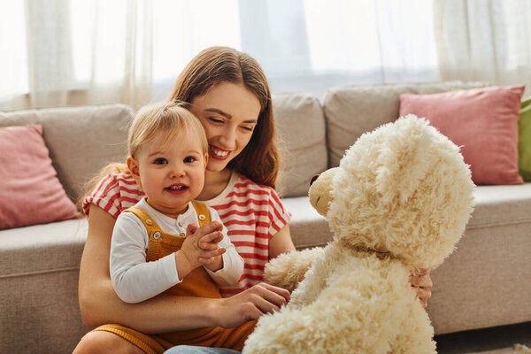Молодая мать с любовью обнимает свою малышку, обе улыбаются, в то время как малыш крепко сжимает плюшевого мишку.