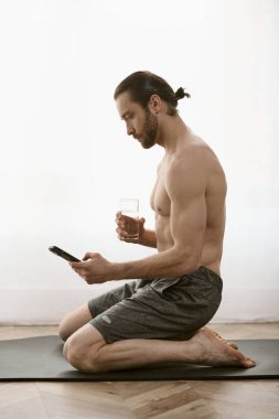 Tişörtsüz bir adam elinde içki ve telefonla mindere oturur..