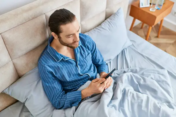 男は平和的にベッドの上に座り 携帯電話の画面に焦点を当てています — ストック写真