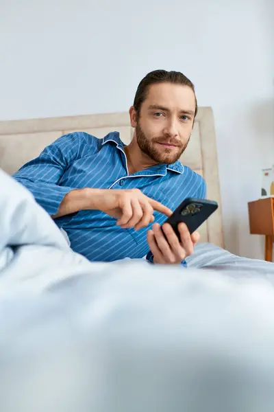 落ち着いたエネルギーに囲まれたベッドに座っている間 携帯電話で従事する男 — ストック写真