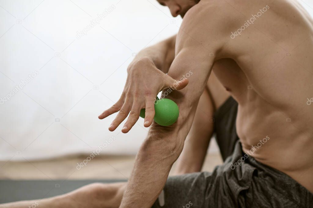 A shirtless man peacefully holds a green massage ball in his right hand while practicing yoga at home.