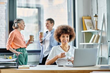Çok çalışan Afro-Amerikan iş kadını dizüstü bilgisayar kullanıyor, arkaplanda çeşitli meslektaşları var..
