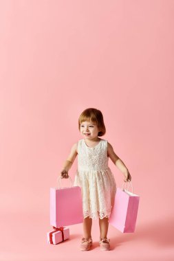 Beyaz elbiseli küçük bir kız pembe arka planda pembe alışveriş torbaları tutuyor..