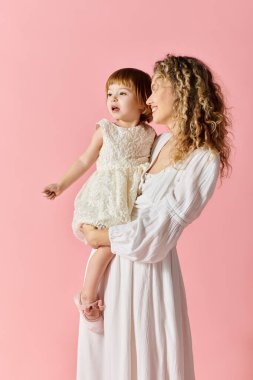 Beyaz elbiseli bir anne pembe arka planda beyaz elbiseli bebeğini sevgiyle kucağına alır..