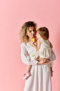 A woman holding a child while eating an apple on a pink background. clipart