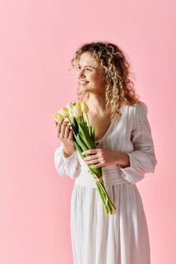 Beyaz elbiseli kadın pembe arka planda laleler tutuyor..