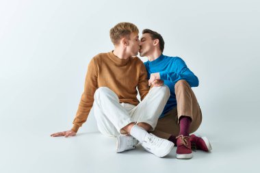 İki genç adam, gündelik giysiler içinde yerde oturmuş öpüşüyorlar..