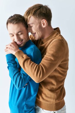 Sıradan giyinmiş iki adam, gri bir zemine karşı sevgi dolu bir kucaklaşmayla sarılıyorlar..