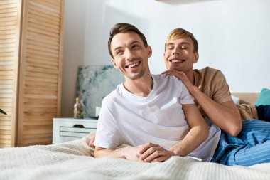 Gündelik giyinmiş iki adam bir yatağın üzerinde otururken birlikte güzel vakit geçiriyorlar..
