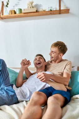 Two men in casual attire sit closely together on a bed. clipart