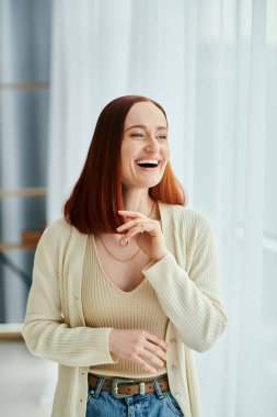 A red-haired woman laughs joyfully in front of a window with the setting sun casting a warm glow. clipart