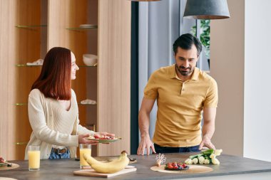 Kızıl saçlı bir kadın ve sakallı bir erkek modern bir mutfakta birlikte yemek pişiriyor. Yemek hazırlama ve kaliteli zaman geçirme konusunda kaynaşıyorlar..