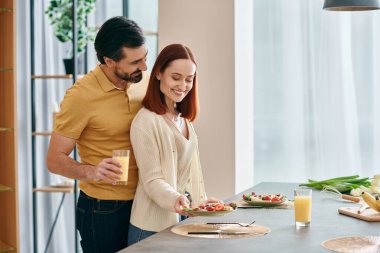 A beautiful adult couple, a redhead woman, and a bearded man are spending quality time together while eating breakfast in a modern kitchen. clipart