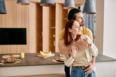 Redheaded woman and bearded man in a warm embrace in a modern kitchen, showing affection and connection. clipart