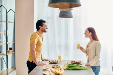 Kızıl saçlı bir kadın ve sakallı bir adam modern dairelerinin mutfak tezgahında birlikte yemek hazırlıyorlar..