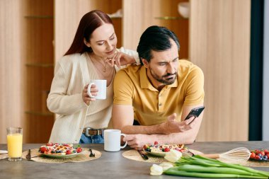 Kızıl saçlı bir kadın ve sakallı bir adam modern bir dairede kahvaltı ederken telefonlarına sarılmış..