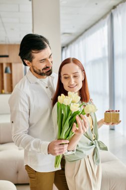 A bearded man lovingly offers a bouquet of tulips to a redhead woman in a modern apartment. clipart