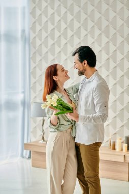 A redhead woman and bearded man stand in a room filled with flowers, enjoying quality time together in a contemporary setting. clipart