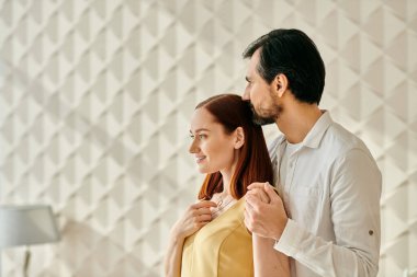 Şık bir yetişkin çift, kızıl saçlı bir kadın ve sakallı bir adam, minimalist beyaz bir duvarın önünde yan yana duruyorlar..