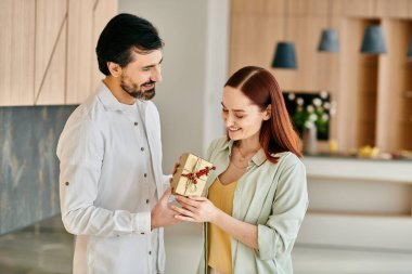 Kızıl saçlı bir kadın ve sakallı bir adam modern bir mutfakta hediye kutusunu paylaşıyorlar, gülümseme ve neşe saçıyorlar..