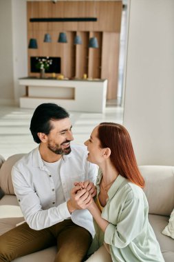 Güzel bir yetişkin çift, kızıl saçlı bir kadın ve sakallı bir adam modern bir dairede rahat bir kanepede birlikte oturuyorlar..