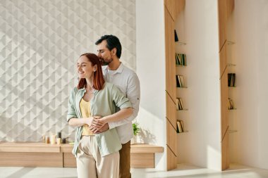 Kızıl saçlı bir kadın ve sakallı bir adam modern bir oturma odasında duruyor, birbirlerine eşlik ederek ve şık bir dekorla eğleniyorlar..