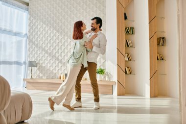 Güzel, kızıl saçlı bir kadın ve sakallı bir adam modern bir apartman dairesinde kıvrılıp dans ediyorlar..