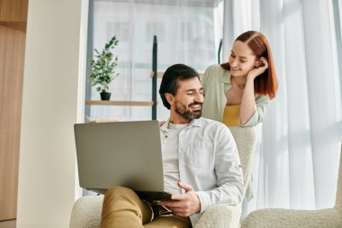 Kızıl saçlı kadın ve sakallı adam birlikte oturuyor, modern apartmandaki dizüstü bilgisayara odaklanmış..