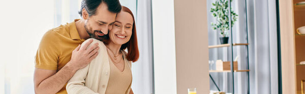 Рыжая женщина и бородатый мужчина обнимаются на современной кухне, олицетворяя любовь и единение..