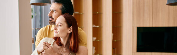 A bearded man and a redhead woman affectionately hug in their stylish living room, radiating warmth and love.