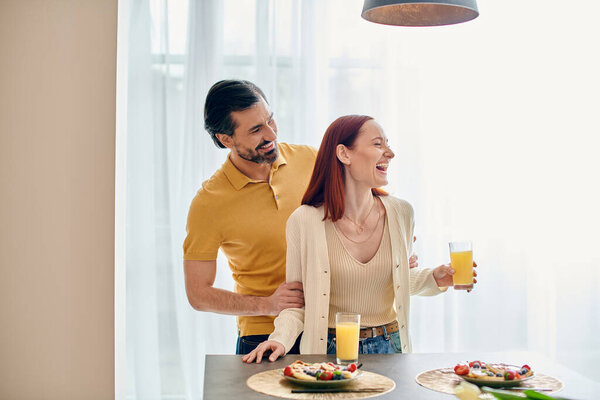 A redhead woman and bearded man enjoy breakfast together in a modern apartment, connecting over food and conversation.