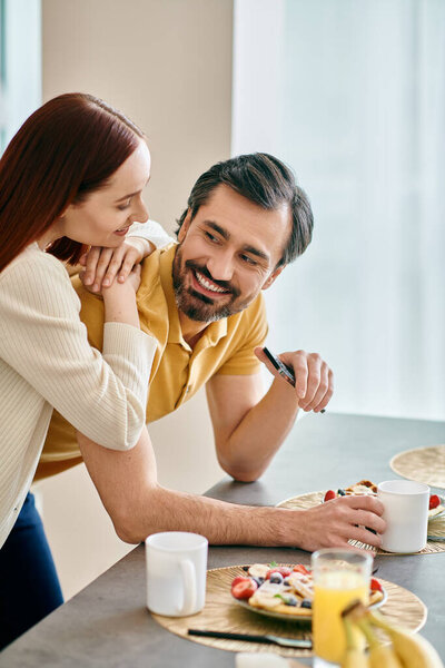 A redhead woman and bearded man share breakfast while hugging in their modern apartment, connecting intimately.