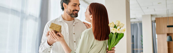 A bearded man in a modern apartment offers a bouquet of flowers to his redhead woman, creating a sweet and romantic moment.