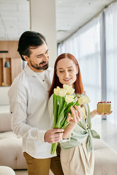 A bearded man lovingly offers a bouquet of tulips to a redhead woman in a modern apartment.
