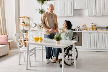 Tekerlekli sandalyedeki engelli bir kadın, evdeki mutfağın sıcak ortamında kocasıyla konuşuyor..