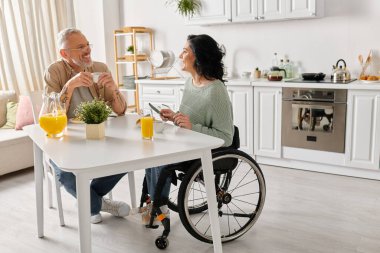 A man in a wheelchair engaged in conversation with a woman in a wheelchair in a cozy kitchen setting at home. clipart