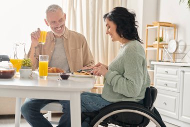 Tekerlekli sandalyede bir adam ve bir kadın evlerinin mutfağında birlikte kahvaltı ediyorlar..