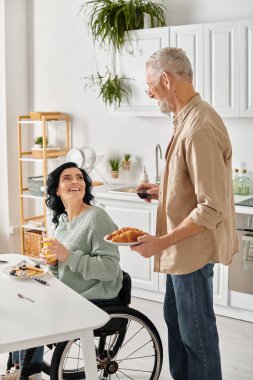 A woman in a wheelchair happily looking at husband holds a plate with croissant in a cozy kitchen setting at home. clipart