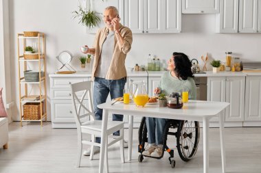 A man stands beside his disabled wife, who is sitting in a wheelchair at a table in their kitchen at home. clipart
